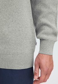 Grauer Strickpullover mit gerippten Bündchen und Saum, strukturiertem Stoff und einem kleinen Markenetikett, das an der Seitennaht sichtbar ist.