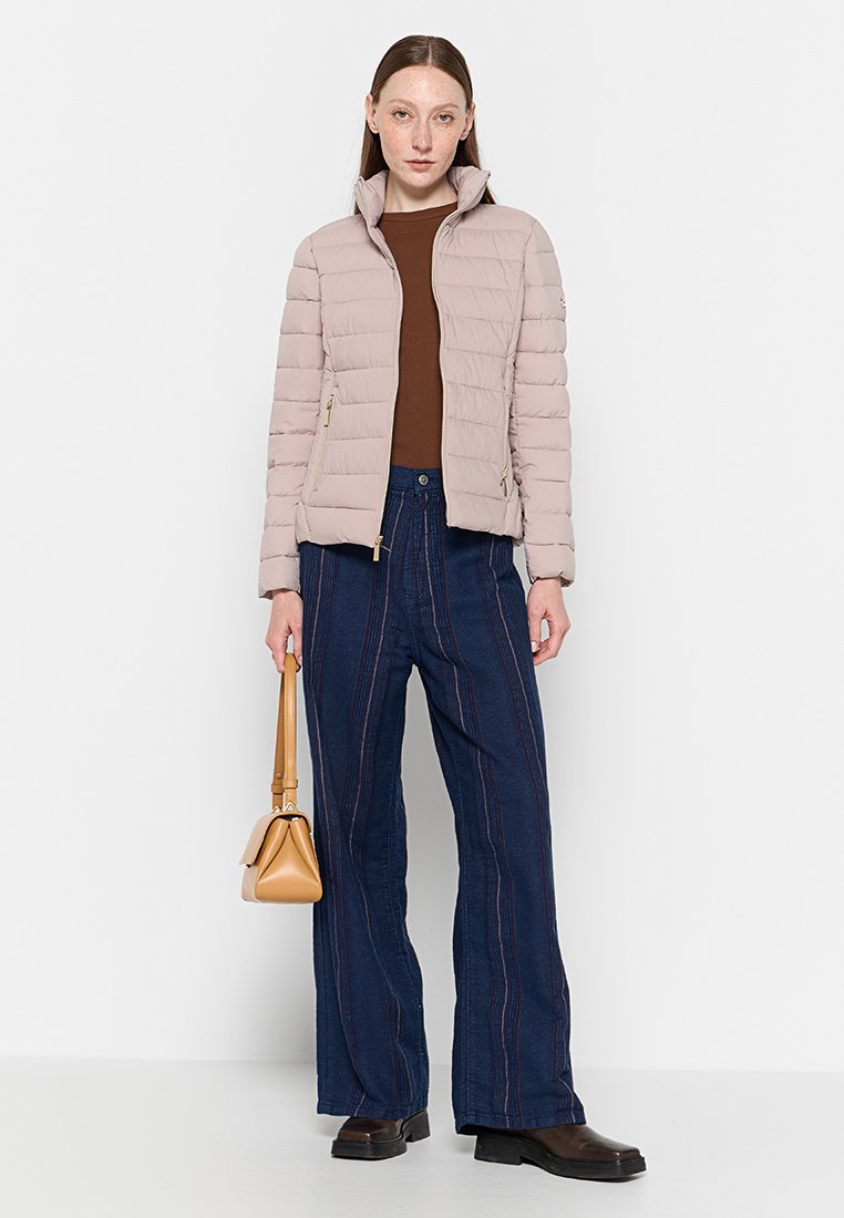 Giacca imbottita rosa chiaro con colletto alto, tasche con zip, abbinata a jeans blu scuro a gamba larga con cuciture verticali e stivali marroni.