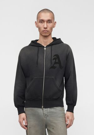 Fiatal férfi fekete cipzáros kapucnis pulóverben, amelynek mellkasán „A” logó látható, és kopott szürke farmerben, egyszínű háttér előtt áll.