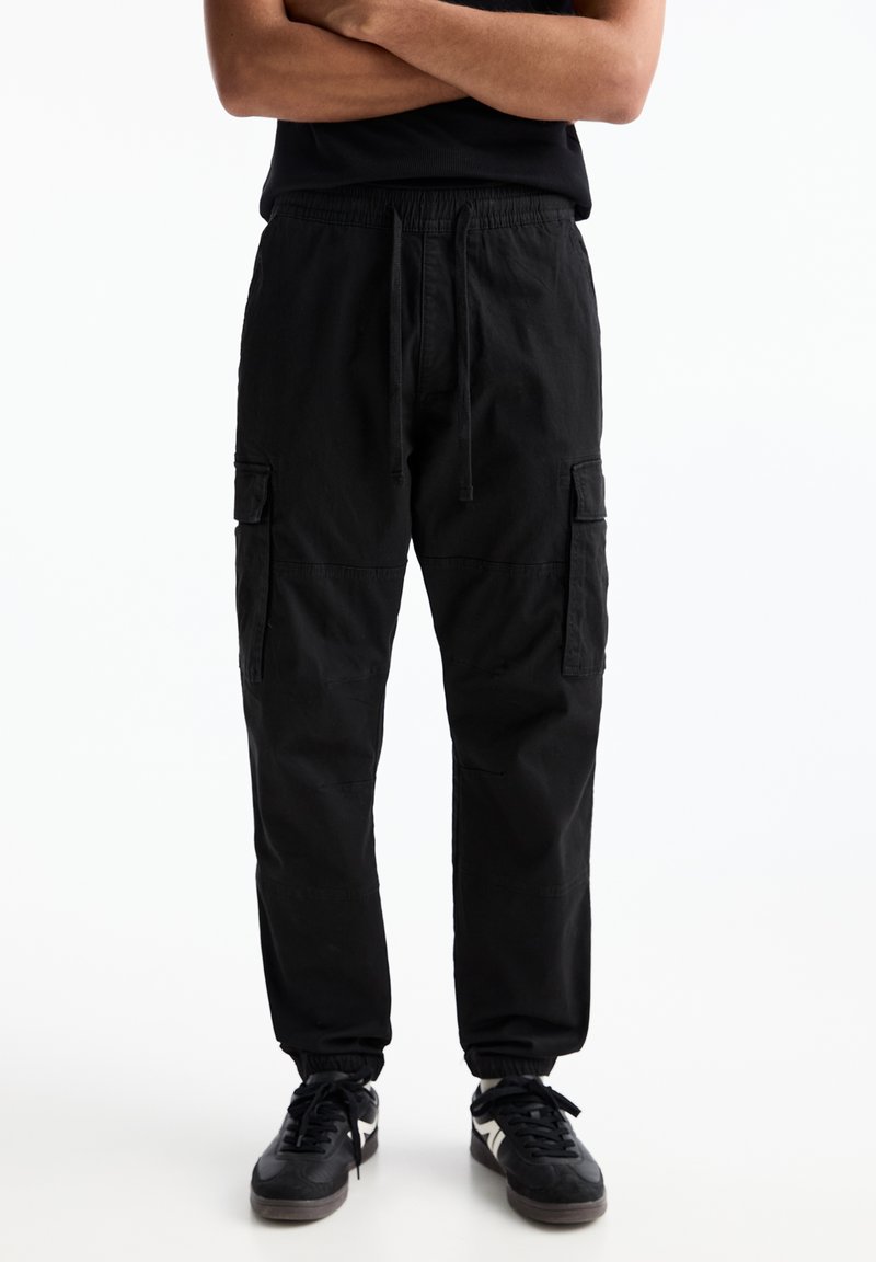 Zwarte cargopants met een drawstring tailleband, voorzien van twee zijzakken en een relaxte pasvorm. Zacht materiaal met elastische manchetten bij de enkels.