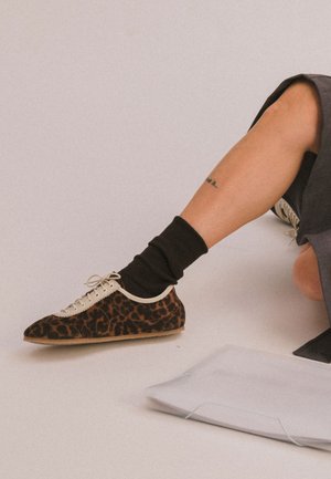 Jambe pliée avec une basket imprimé léopard, chaussette noire, tatouage sur le bas de la jambe et jupe grise ; document blanc plié sur le sol à côté de la jambe.