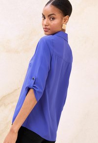 Kraljevsko modra bluza z ovratnikom, 3/4 zavihanimi rokavi, pritrjenimi z gumbnimi trakovi, in ukrivljenim robom. Gladek tekstura in lahka tkanina.