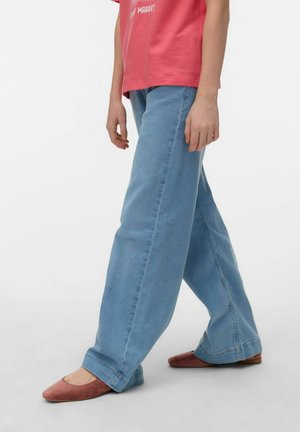 Személy világoskék, bőszárú farmernadrágot, rózsaszín pólót és barna lapos cipőt visel, oldalirányban áll egy sima fehér háttéren.