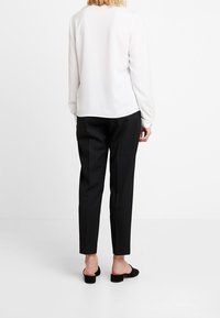 Nainen seisoo selkä kameraan päin valkoisessa pitkähihaisessa paidassa, mustissa kapeissa housuissa ja mustissa avonaisissa kengissä valkoista taustaa vasten.