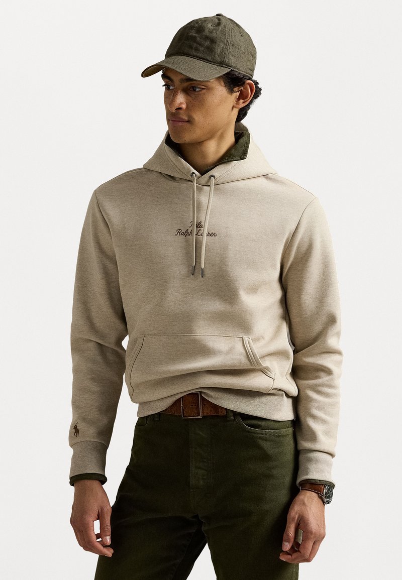 Μπεζ φούτερ με καπροσκέπαστο και κορδόνια στο καπέλο, που διαθέτει κεντητό λογότυπο. Συνδυασμένο με σκούρα πράσινα παντελόνια και καφέ ζώνη. Καθημερινό καπέλο.