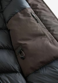 Padded Jacke aus einer Kombination von schwarzem und dunkelbraunem Material, mit einer Reißverschlusstasche und verstellbarem Kordelzug an der Seite.