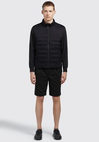 Sort polstret vest med lynlås og høj krave, parret med sorte shorts og sorte sneakers. Glat tekstur og minimalistisk design.
