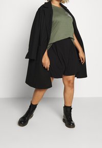 Overmaatse zwarte jas, olijfgroen T-shirt met logo, zwarte shorts en zwarte gevechtslaarzen. Kenmerkt zich door een relaxte pasvorm en zachte texturen.