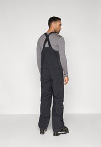 Μαύρη στολή για σκι με φαρδιές τιράντες, άνετη εφαρμογή και πλαϊνές τσέπες. Κατασκευασμένη από ανθεκτικό, ανθεκτικό στο κλίμα υλικό.