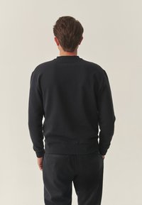 Czarna bluza z okrągłym dekoltem, ściągaczami na rękawach i u dołu. Miękka faktura, luźny krój i minimalistyczne detale, widziana od tyłu.