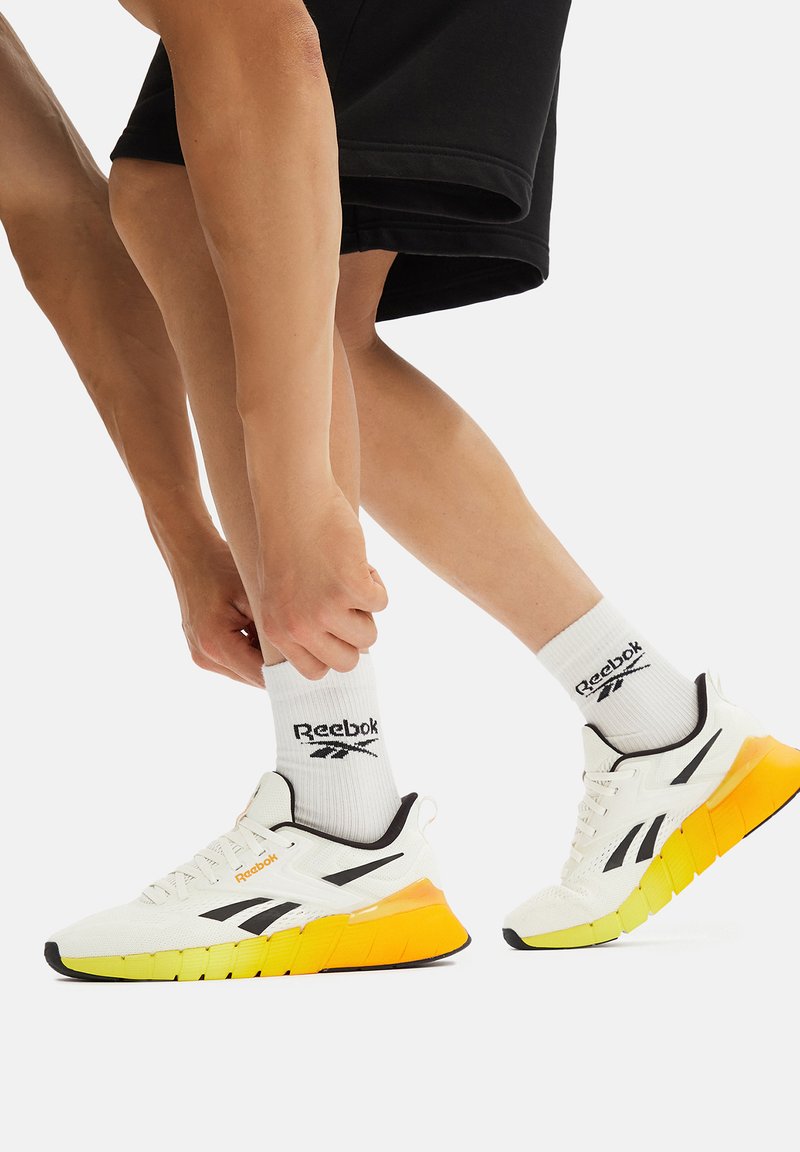 Osoba ubrana w białe trampki Reebok z żółtymi podeszwami i białe skarpety Reebok, poprawiająca skarpetę na jednej nodze na jednolitym tle.