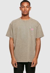 Camiseta de algodón de manga corta en color marrón claro con un pequeño gráfico de flamenco rosa en el lado izquierdo. Cuello redondeado, corte relajado y detalles de costura.