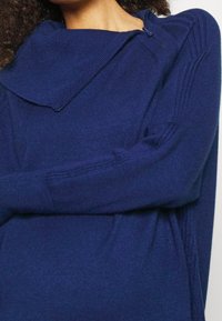 Maglione oversize blu navy con collo a cowl e dettagli a costine diagonali sulle maniche. Il materiale sembra morbido e texture.