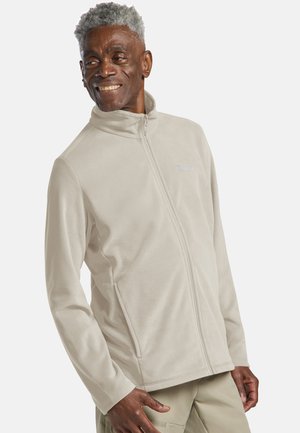 Lächelnder Mann, der eine hellbeige Fleecejacke mit Reißverschluss und beige Hose trägt, steht vor einem einfarbigen hellen Hintergrund.
