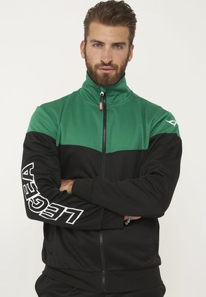 Uomo con barba che indossa una giacca verde e nera con zip e marchio "LEGEA" sulla manica, braccia incrociate, guarda la fotocamera su sfondo bianco.