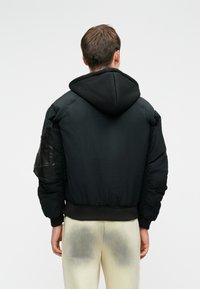 Fekete steppelt dzseki kapucnival, a bal karon textúrázott fekete bőrpólóval, bordázott mandzsettákkal és aljjal.