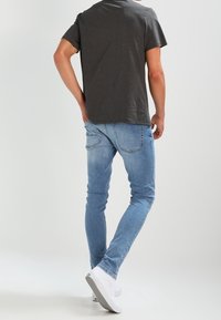 Camiseta gris con manchas combinada con jeans ajustados de color azul claro y zapatillas blancas, vista desde la parte de atrás sobre un fondo claro.