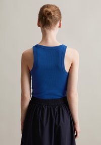 Niebieski prążkowany top na ramiączkach z grubymi paskami, zestawiony z czarną spódnicą na elastycznej talii. Modelka widoczna jest od tyłu.