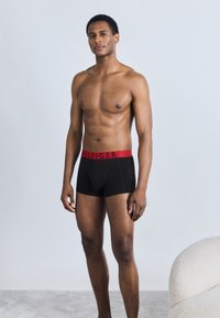 Μαύρα μποξεράκια με κόκκινη ζώνη μέσης και με λογότυπο. Το μοντέλο έχει γυμνασμένο σώμα και στέκεται μπροστά σε ανοιχτό φόντο.