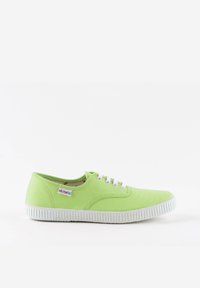 Zapatilla de lona verde con suela de goma blanca y ojales, con una etiqueta de logo en el lateral. Punta redondeada y diseño de corte bajo.