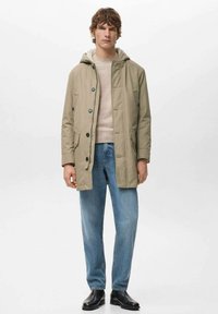 Chaqueta con capucha de color beige con cierre de botones, dos bolsillos frontales y un interior suave, combinada con jeans azul claro y botas negras hasta el tobillo.