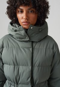 Giacca a puffo di un verde tenue, con colletto alto, chiusure a bottoni e vita regolabile per una silhouette strutturata.
