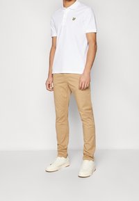 Polo blanc à manches courtes, avec un petit logo jaune ; associé à un chino beige et des baskets blanches. Design simple et épuré.