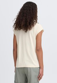 T-shirt beige clair à manches courtes avec une coupe décontractée, en tissu doux. Dispose d'un détail de couture dans le dos et d'un ourlet arrondi, associé à un short vert.