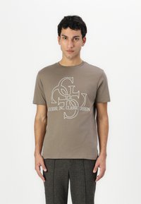 Brown cotton t-shirt with a round neckline, featuring a white logo print of "GUESS?, INC. CLASSIC DESIGN" at the center.