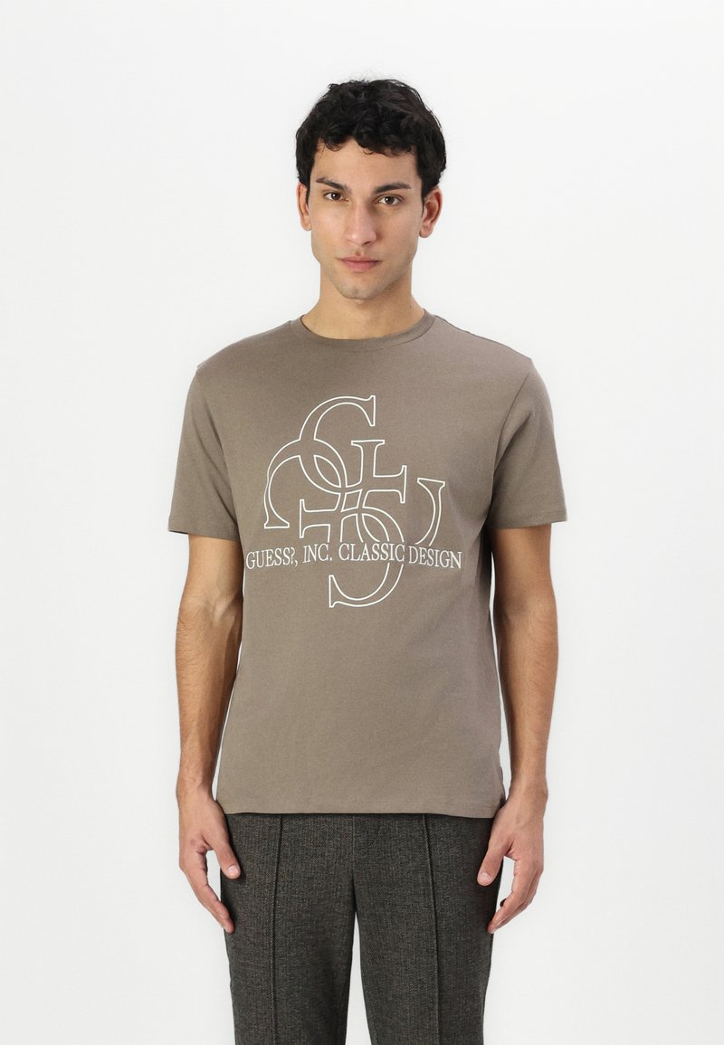 Brown cotton t-shirt with a round neckline, featuring a white logo print of "GUESS?, INC. CLASSIC DESIGN" at the center.
