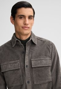 Camicia giacca in corduroy grigio con due tasche sul petto, chiusura a bottoni e texture a coste; indossata sopra un top verde scuro a collo alto.