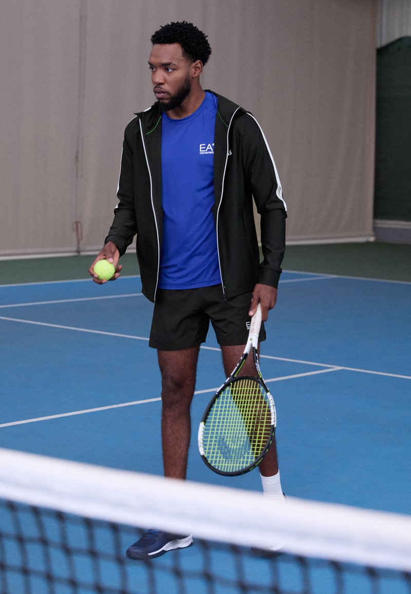 Schwarze Sportjacke mit weißen Akzenten, blaues Sporthemd, schwarze Shorts und Tennisschuhe. Hält einen Tennisball und einen Schläger auf einem Platz.