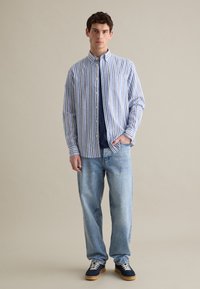 Camicia a righe blu e bianca, sovrapposta a una maglietta blu navy, abbinata a jeans azzurri e scarpe da ginnastica navy.