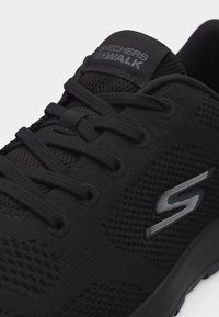 Černá teniska Skechers GOwalk s textilním síťovaným materiálem, zavázanými tkaničkami a stříbrným logem „S“ na boku.