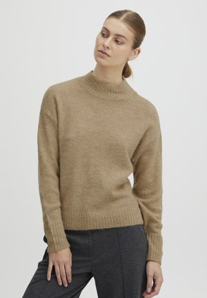 Beige knit sweater with a high collar and ribbed cuffs, featuring a soft texture and slightly boxy fit, paired with dark gray trousers.
