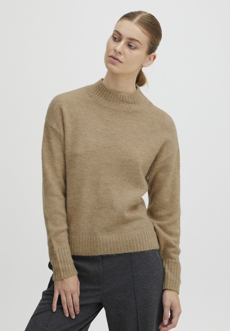 Pull en tricot beige avec un col montant et des poignets côtelés, présentant une texture douce et une coupe légèrement boxy, associé à un pantalon gris foncé.
