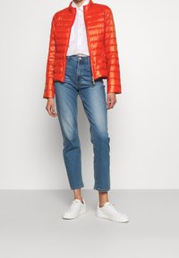 Giacca trapuntata rossa con colletto alto, chiusura zip e texture liscia; abbinata a jeans in denim azzurri e sneaker bianche.