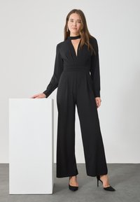 Combinaison noire à manches longues, décolleté en V profond et détails à la taille. Tissu lisse, jambes larges, accompagnée de talons hauts noirs.