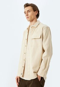 Camisa de botón color crema con un bolsillo frontal, hecha de una tela suave. Presenta botones en tono dorado y un ajuste relajado.