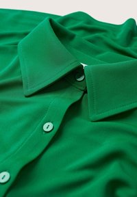 Polo vert avec une texture lisse, col classique et boutons clairs, présentant un design uni et un tissu plié.