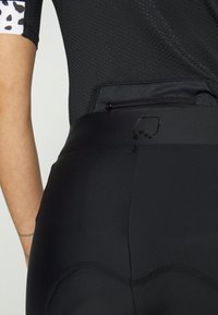 Calções de ciclismo pretos com uma textura suave, apresentando um bolso com fecho nas costas e um acabamento em padrão contrastante nas mangas.