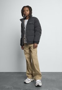 Barn med dreadlocks iført en svart dunjakke, hvit skjorte, beige bukser og hvite sneakers står mot en ensfarget grå bakgrunn.