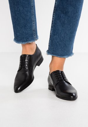 Black leather lace-up oxfords worn with blue frayed-hem jeans on a white floor background.