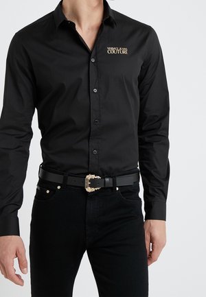 Zwart overhemd met knopen en het logo "Versace Jeans Couture" en een zwarte riem met een rijkelijk versierd gouden gesp, gedragen met een zwarte broek.