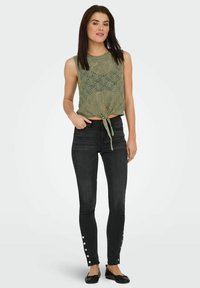 Top senza maniche di colore verde oliva, lavorato a maglia con un intricato motivo in pizzo e un nodo sul davanti. Abbinato a jeans skinny neri con dettagli a bottoni a pressione.