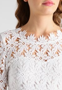 Hvid blonde top med indviklede blomster mønstre, en scallop-udskæring og teksturerede detaljer, der viser delikat håndværk og design.