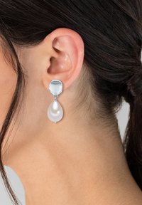 Strieborné visiace náušnice majú štruktúrovaný okrúhly vrch a hladký oválny biely perlový prívesok, ktorý pridáva eleganciu a kontrast.