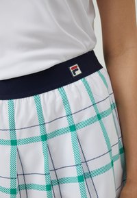 Sezione vita di una gonna sportiva bianca con motivo a scacchi blu e verde, elastico in vita color navy e una piccola etichetta con logo sulla cintura.