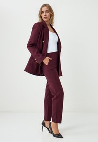 Bordeaux op maat gemaakte blazer met gouden knopen, gecombineerd met bijpassende slim-fit broek en een witte top. Zwarte hoge hakken maken de look compleet.