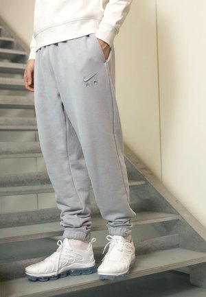 Osoba nosící světle šedé tepláky Nike Air a bílé sportovní boty stojí na šedých vnitřních schodech.